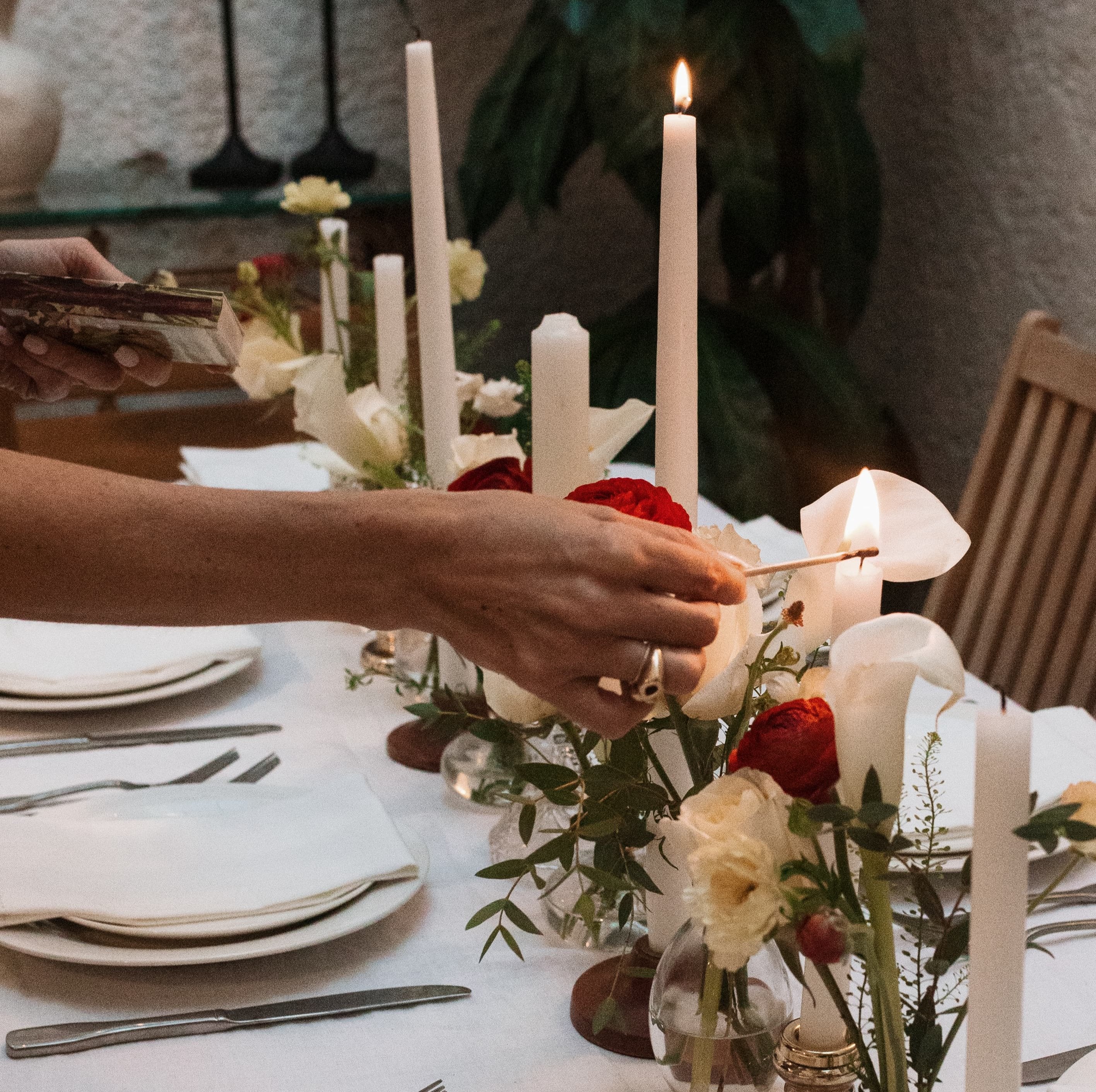 Hand holding a match lighting a tall candle on a preset dining table. 