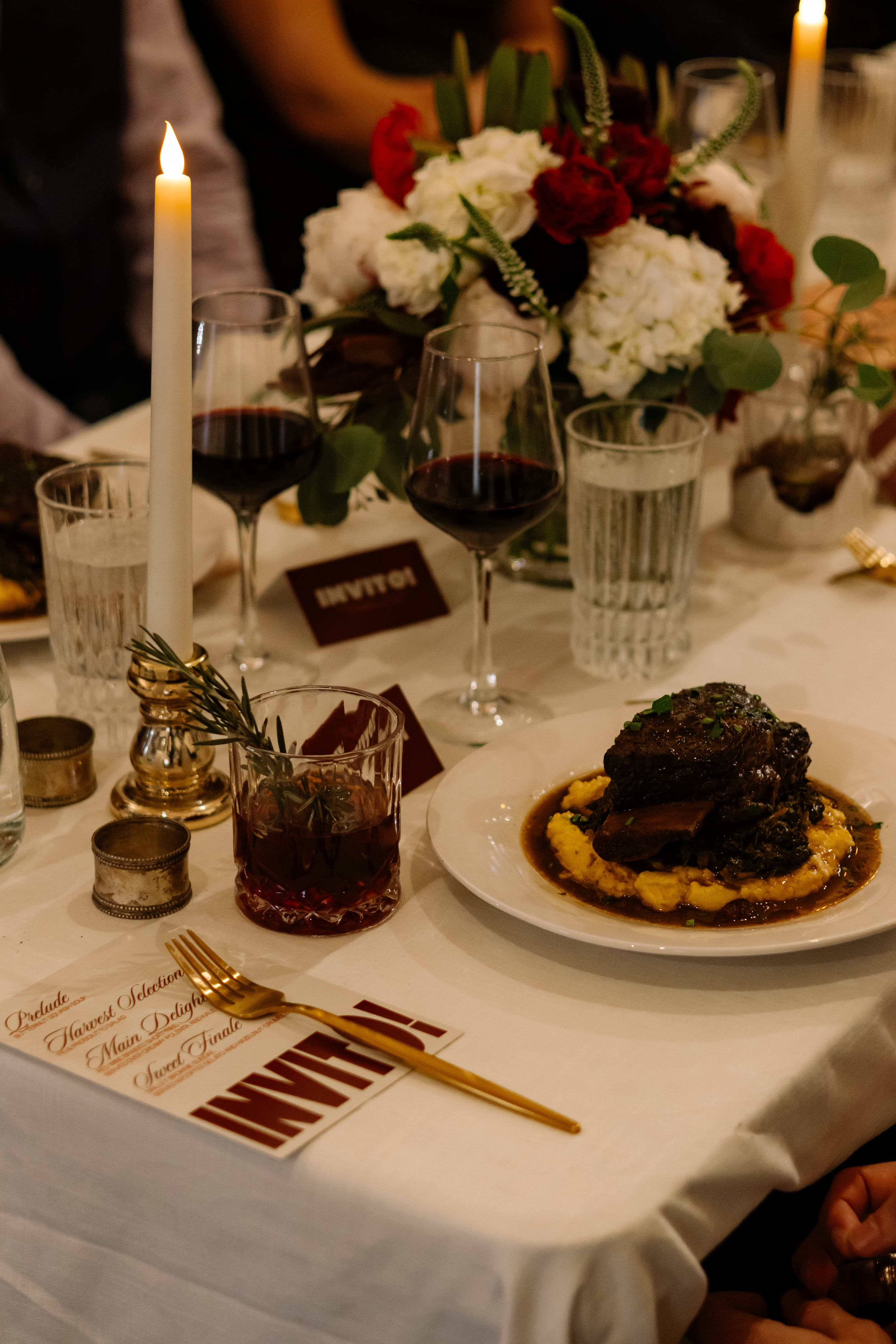 An elegantly set dinner table featuring a plate of gourmet short ribs on mashed potatoes, surrounded by glasses of red wine, water, and a rosemary-garnished cocktail. The table is adorned with a floral centerpiece of red and white blooms, lit candles, and a printed menu card displaying the Invito logo.